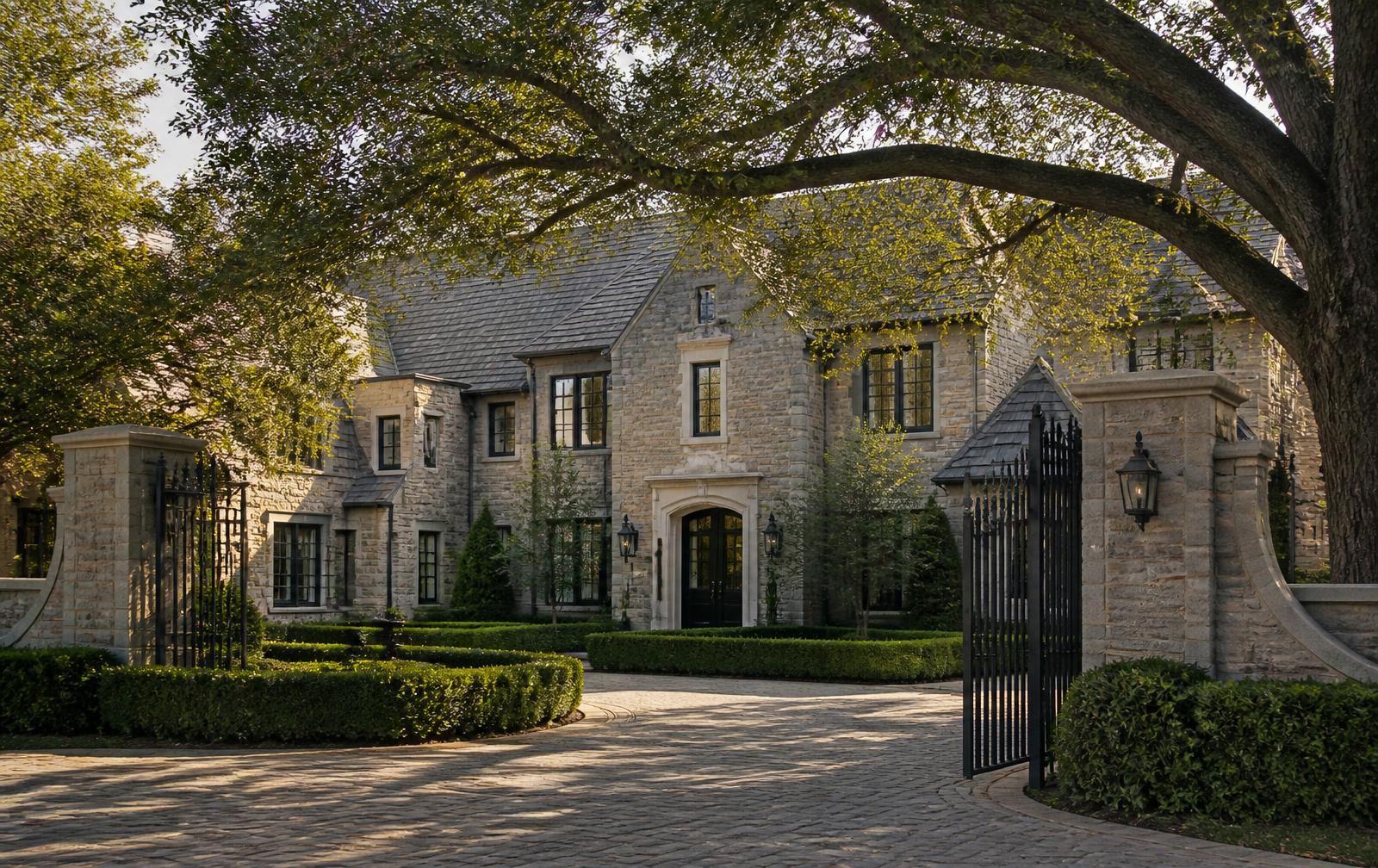 Stone-and-slate European-style Charlotte estate behind an open wrought-iron gate under mature oaks at golden hour